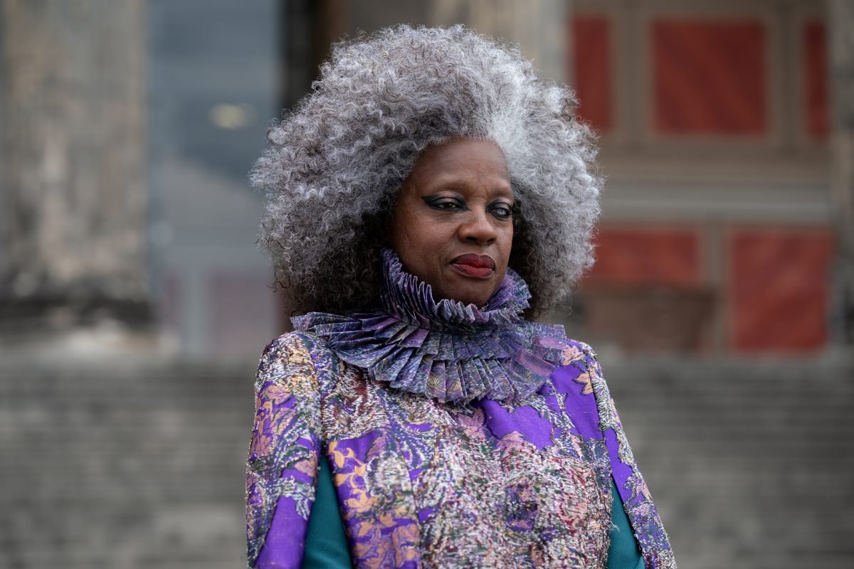 Dr. Volumnia Gaul stands in front of stairs, wearing a purple-patterned top