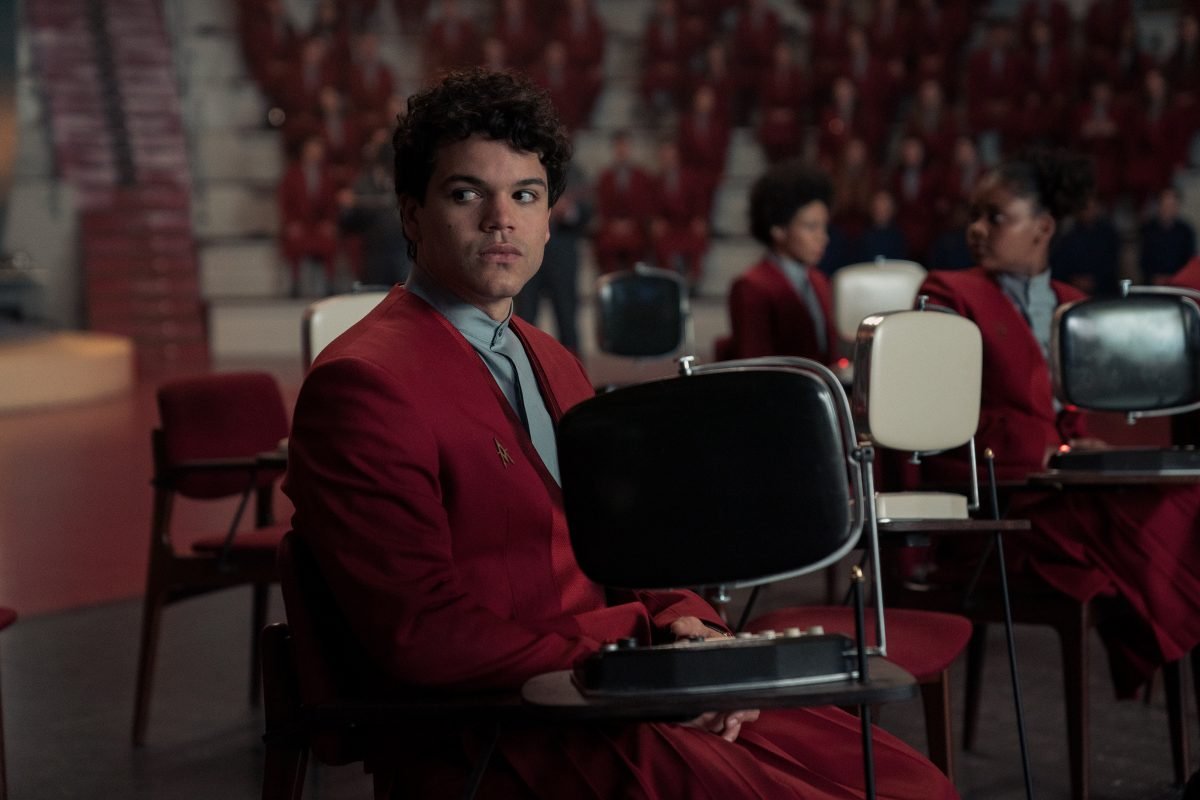Sejanus Plinth, wearing a red school uniform, sits at a black desk beside other students