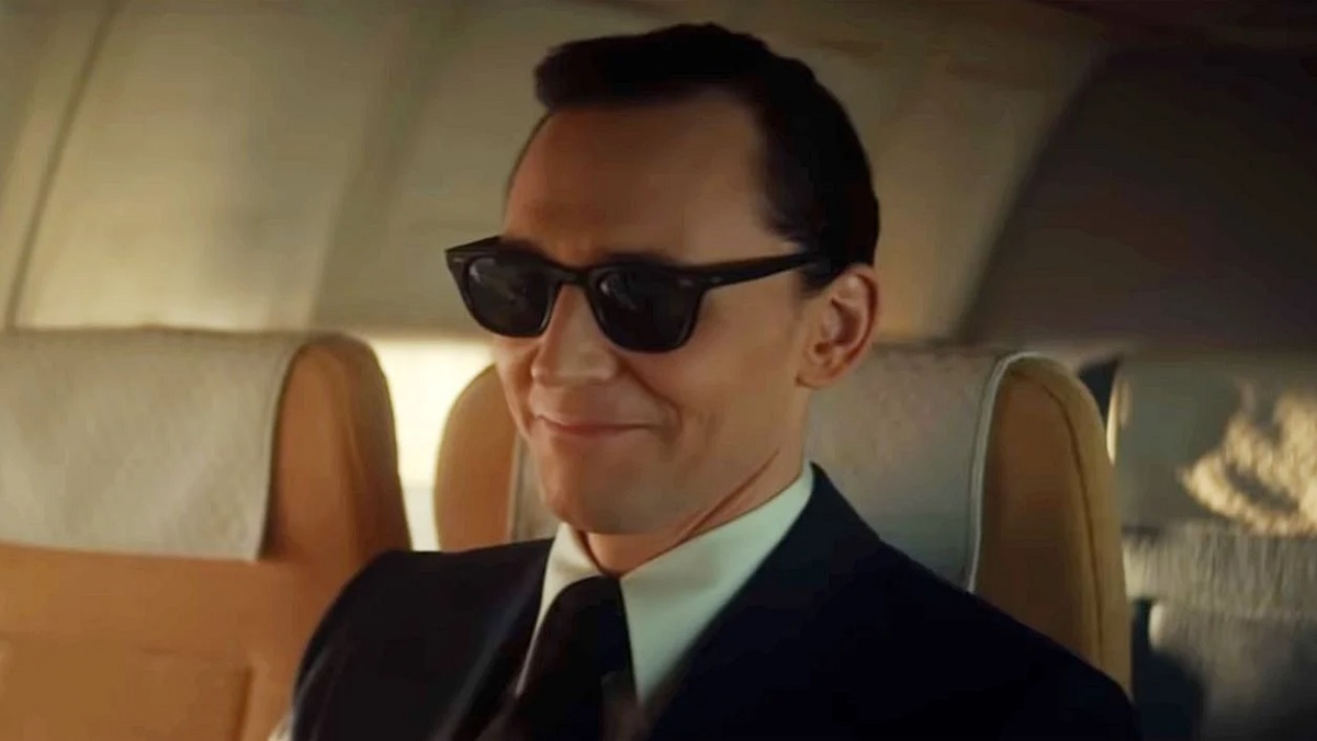 Tom Hiddleston as Loki dressed in a snappy suit, slicked hair, and sunglasses aboard a 1971 airplane assaying the infamous hijacker D.B. Cooper.